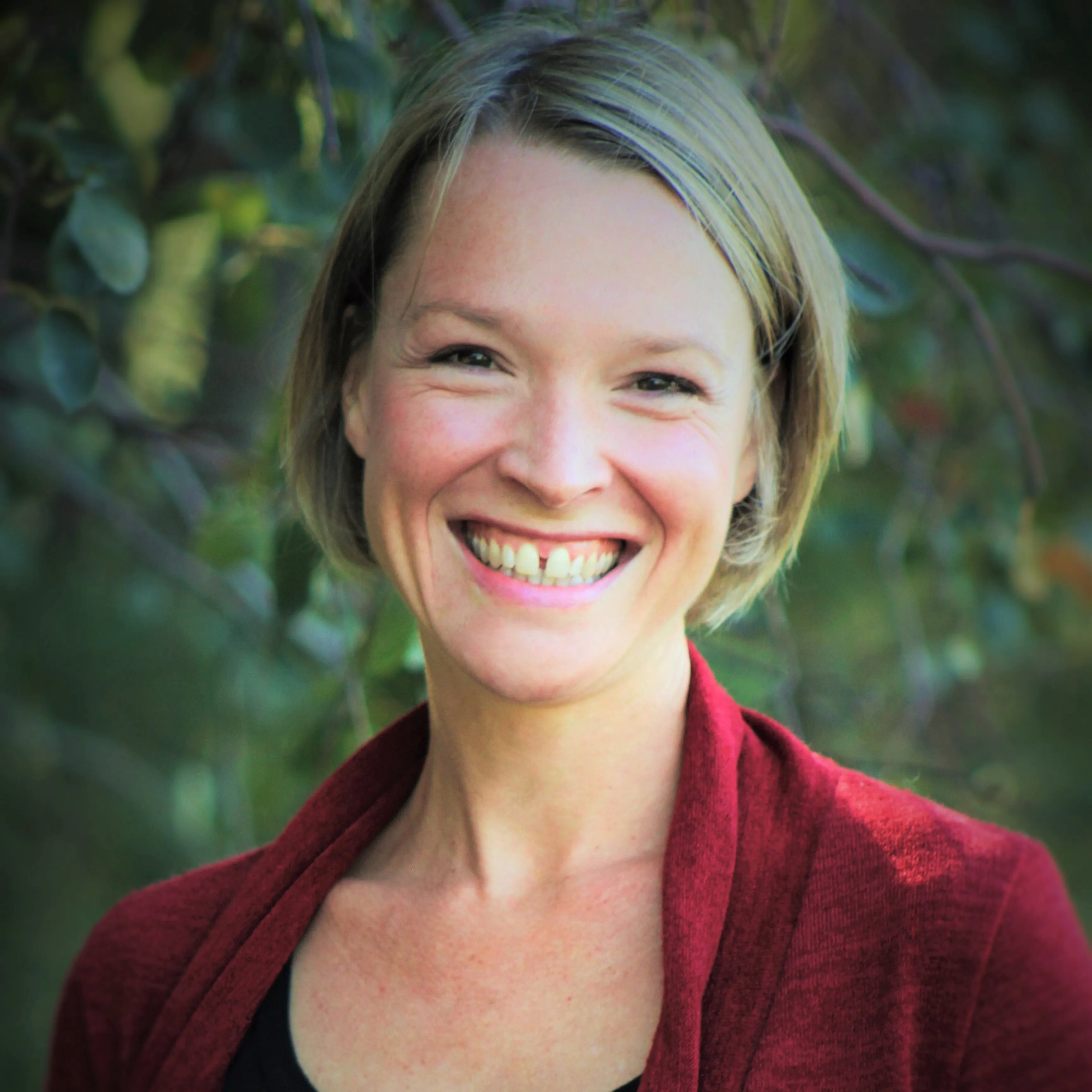 Smiling woman with short blonde hair wearing a red cardigan and black top, standing outdoors with green foliage background.